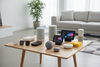 “A premium smart home setup displayed on a clean modern table in a bright, minimal living room. High-quality, realistic product arrangement featuring smart assistants and hubs in perfect condition: Amazon Echo Dot, Echo Show, Echo Studio, Google Nest Mini, Google Nest Hub, Google Nest Hub Max, Apple HomePod Mini, Apple HomePod, Xiaomi XiaoAI Speaker, Tencent Tingting Speaker, Samsung SmartThings Hub, Hubitat Elevation Hub, Aqara Hub M2 and E1, Home Assistant Yellow, Homey Pro Hub, Tuya/Smart Life Zigbee Gat
